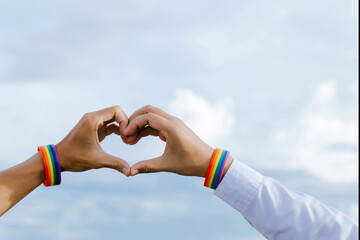 Closeup shot of a gay couple holding hands with a rainbow wristband made hands forming a heart shape in the sky, Concept of LGBT, activism, community, homosexuality, and freedom.
