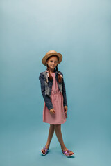 Fashionable girl with red hair in pink dress, straw hat and denim jacket looking into camera on isolated blue background..