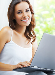 Portrait image of beautiful happy smiling brunette woman using laptop, sitting against window. Working at home, on distance concept.