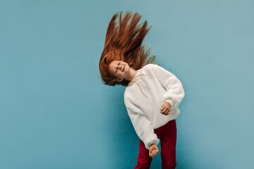 Lovely girl with freckles in modern trousers and white warm sweater smiling and playing her foxy long hair on blue backdrop..