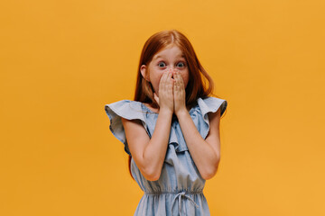 Shocked open-eyed child in denim stylish dress covers mouth and looks into camera in surprise on...