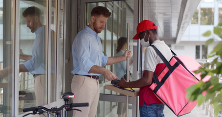 African courier in safety mask handing fresh food to young businessman customer