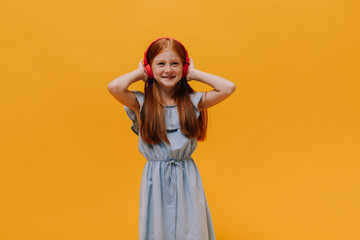 Happy redhead little girl listens to music in massive red headphones and smiles sincerely on isolated orange background.