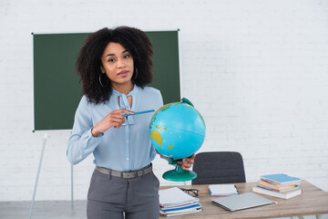 African american teacher pointing at globe and looking at camera in classroom
