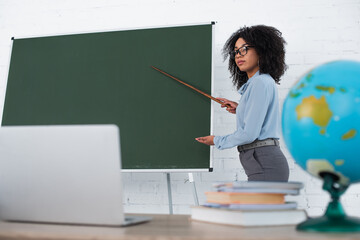 African american teacher pointing at chalkboard near blurred laptop during online lesson in school