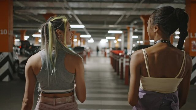 Back view of two diverse trendy ladies with unusual appearance talking together, walking at underground parking