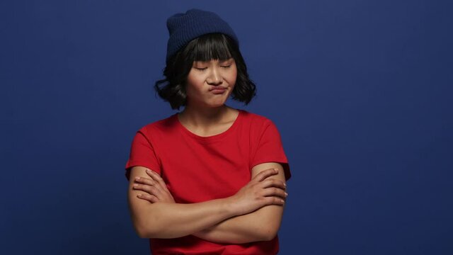 A disappointed angry asian woman is holding her hands while looking to the side standing isolated over blue wall in the studio