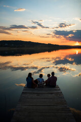 Naklejka na ściany i meble family of four people sitting on the shore on a wooden bridge of a large lake in summer and watching the beautiful sunset with dog back view
