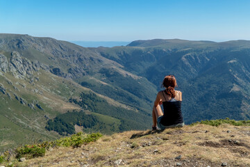 Naklejka na ściany i meble A woman sitting on top of a mountain in a relaxing mood and looking at a beautiful view of peaks, forests and blue skies