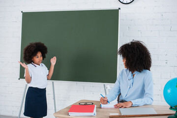 African american pupil showing shrug gesture near teacher and chalkboard