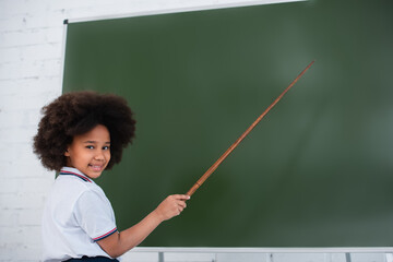 Smiling african american schoolkid looking at camera and pointing at chalkboard in classroom