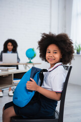 Smiling african american kid holding backpack in classroom
