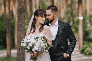 A bearded, stylish groom in a suit and a beautiful bride in a white dress with a bouquet in their hands outdoors in the park tenderly hug. Wedding portrait of newlyweds in love.