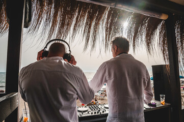 Back view of disc jockeys playing music for tourist people at club party outdoors on the beach -...