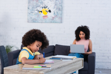 African american kid doing schoolwork near blurred mother with laptop