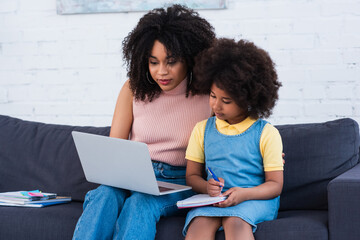 Obraz na płótnie Canvas African american mother holding laptop while daughter writing on notebook during online lesson