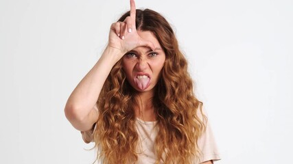 A close-up view of an emotional woman is doing loser gesture standing isolated over a white wall in the studio
