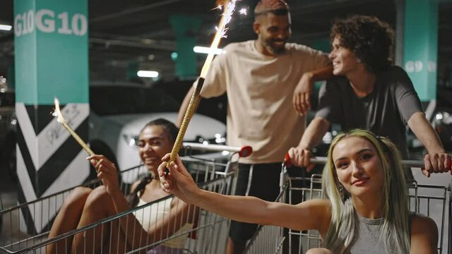 Group portrait of young people laughing together at underground parking, women with fireworks sitting in shopping carts