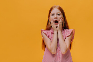 Surprised emotional child looks into camera. Redhead little girl in striped shirt opens mouth and touches cheeks. Girl poses on orange background.