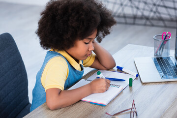 Side view of african american kid writing on notebook near laptop at home