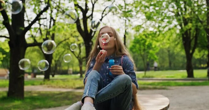 Preteen girl 10 years old blowing soap bubbles at camera outdoors. Child sitting on the bench in the city park. Gimbal shot. Slow motion