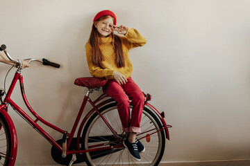 Modern girl with long hair in red pants, sweater cap and black sneakers smiling, showing peace sign and sitting on bicycle on white backdrop..