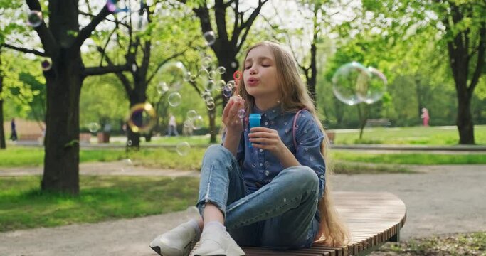 Preteen girl 10 years old blowing soap bubbles outdoors at camera. Child sitting on the bench in the city park. Gimbal shot. Slow motion
