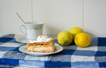 Pastel de hojaldre dulce relleno de crema, acompañado de una taza de café y limones sobre un mantel a cuadros azul.
