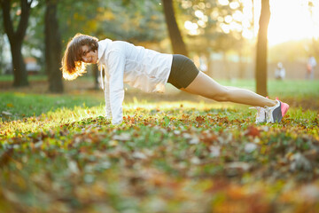 Strong young lady in activewear doing plank exercise at city sunny park. Slim happy brunette...