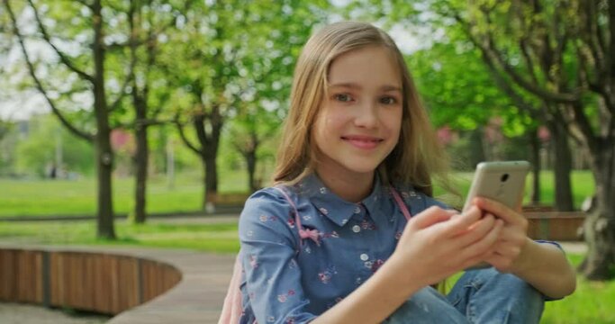 Schoolgirl using cell phone outdoors and smiling at camera. Preteen girl 10 years old sitting on the bench in the city park. Gimbal shot. Slow motion