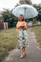 Cheerful young woman in bright clothes under a transparent umbrella in bad weather in the city park