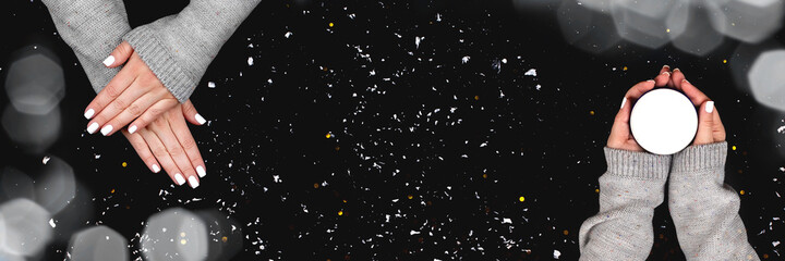 Female hands in gray knitted sweater with white nails manicure with jar with cream on black background with blurred lights bokeh and snow. Panoramic holiday Christmas banner with copy space