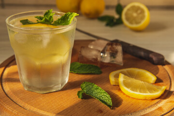 A glass of cold water with lemon and mint on a wooden kitchen board in the morning sun. Melted ice cubes.