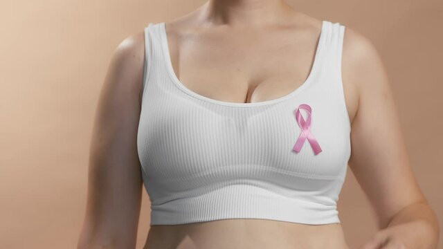 Busty girl in a seamless white bra put a pink ribbon sign on her chest to raise awareness and support women fighting breast cancer. Anonymous slow motion studio shot video on beige background.