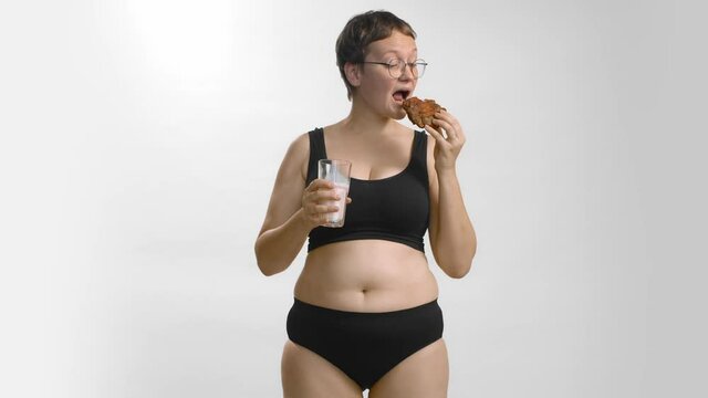 Young beautiful caucasian plus size model in black undewear and glasses with short hair feeling pleasure biting a cookie and drinking cow milk. Studio shot slow-motion high quality video.