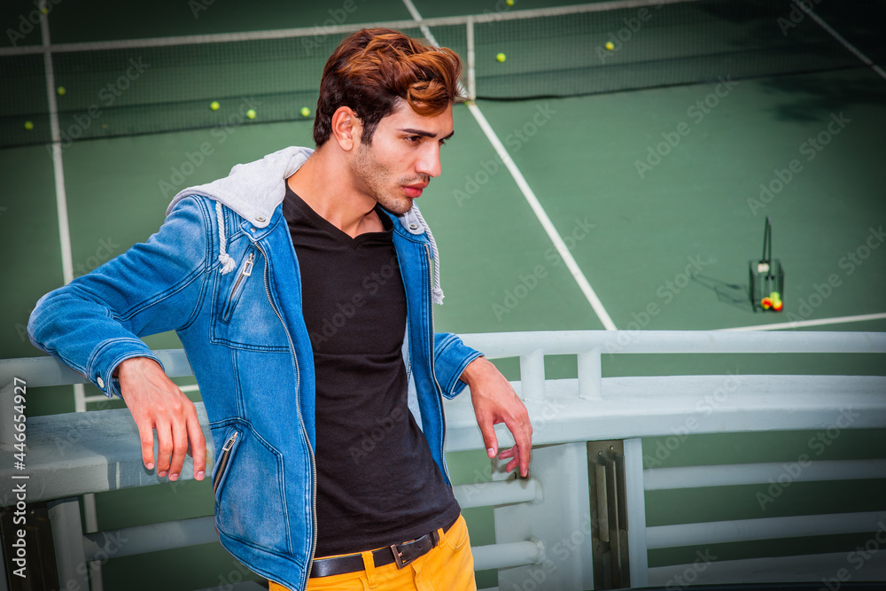 Wall mural Young Man Thinking Outdoors Dressing in a blue jacket with a hood, black underwear, yellow pants, a young handsome Man is standing by a tennis court, arms resting on railings, sad, thinking..