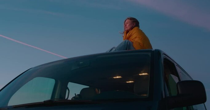 Cinematic shot of young woman cosy cuddled in yellow sleeping bag or blanket, sit on top of camper van. Young female traveller watch sunset from roof or camping van. Wanderlust van life concept