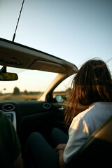 Rearview of an attractive brunette woman riding in the convertible car. Concept of summer adventures and new experiences. Strong sunlight during warm and long summer evenings.