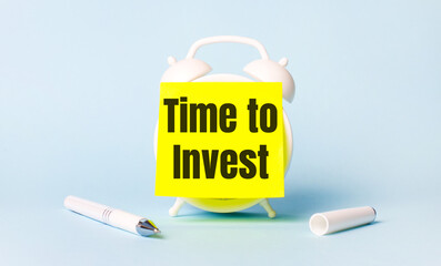 On a light blue background - a white handle and an alarm clock with a bright yellow sticker glued to it with the text TIME TO INVEST