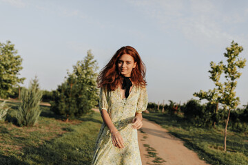 Fototapeta premium Fashionable girl with red long hair and black bow-knit on neck in summer stylish green clothes looking into camera outdoor