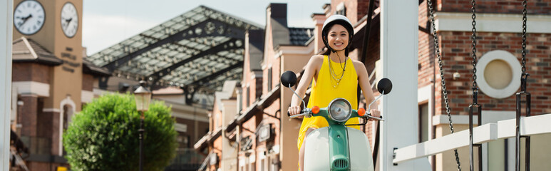 happy asian woman in hardhat enjoying riding scooter on urban street, banner