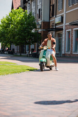 Fototapeta premium pleased african american man on scooter near buildings on urban street