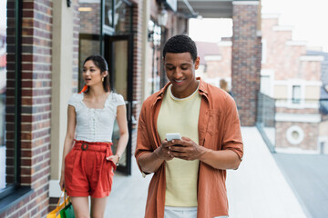 cheerful african american man using mobile phone near asian girlfriend with shopping bags