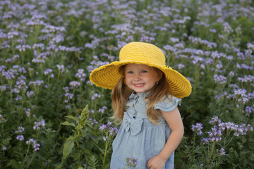 a beautiful happy little smiling blonde girl with ponytails and blue eyes with a yellow hat on her head and a blue muslin dress is standing in a field with beautiful purple flowers