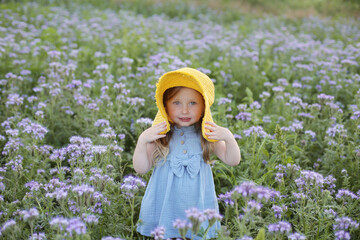 a beautiful happy little smiling blonde girl with ponytails and blue eyes with a yellow hat on her head and a blue muslin dress is standing in a field with beautiful purple flowers