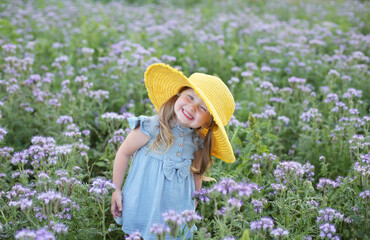 a beautiful happy little smiling blonde girl with ponytails and blue eyes with a yellow hat on her head and a blue muslin dress is standing in a field with beautiful purple flowers