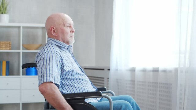 Disabled old man is sitting in a wheelchair at home alone. A handicapped person in a wheelchair is looking out the window. Paralysis and disability.