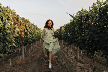 Full length photo of stylish red-haired girl with black bandage on neck in green summer dress walking on background of vineyards