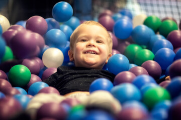 Blonde little boy lying on multi coloured plastic balls in big dry paddling pool in playing centre. Smiling at camera