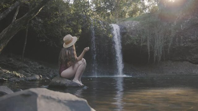 Wide shot of a young woman in swimwear sitting on a rock in front of a waterfall